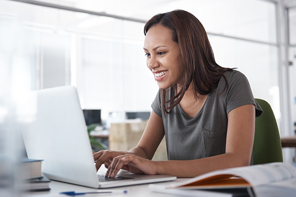 Smiling woman sitting at a desk, leaning forward and typing on laptop computer